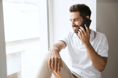 Pencerenin yanında gülümseyen mutlu genç adam cep telefonuyla konuşuyor..