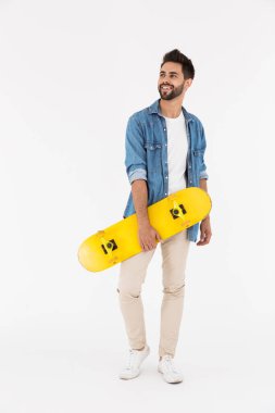 Traşsız, mutlu, gülümseyen ve kaykayla ayakta duran bir adamın tam boy görüntüsü.