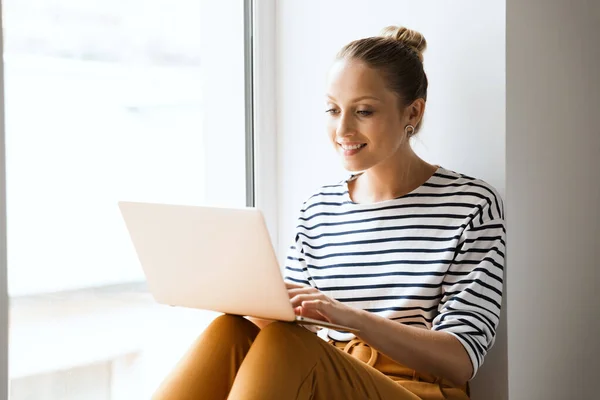 Young happy cheery woman sit near window indoors using laptop computer. — Stock Photo, Image