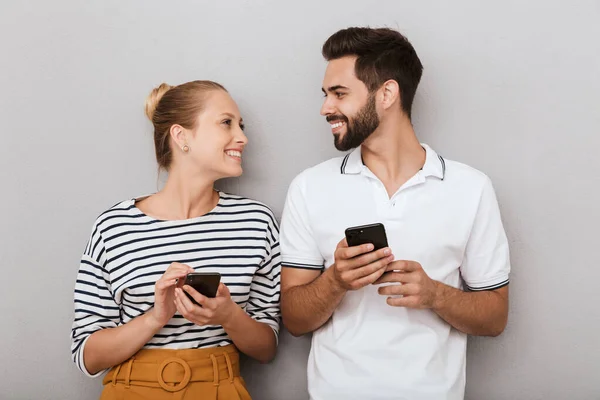 Blij jong positief liefdevol paar vrienden man en vrouw poseren binnen geïsoleerd over grijze achtergrond met behulp van mobiele telefoons. — Stockfoto