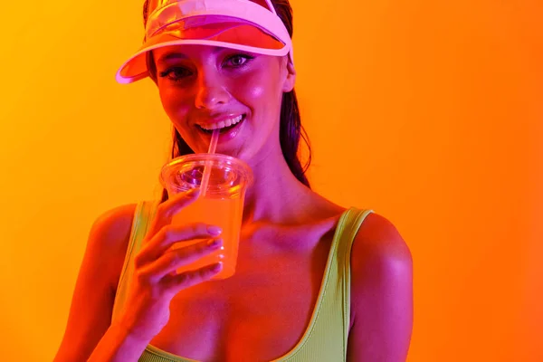 穿着运动服，头戴透明帽帽，穿着运动生活方式的、情绪乐观的时尚女孩，喝着橙红色背景下与霓虹灯隔离的果汁鸡尾酒. — 图库照片