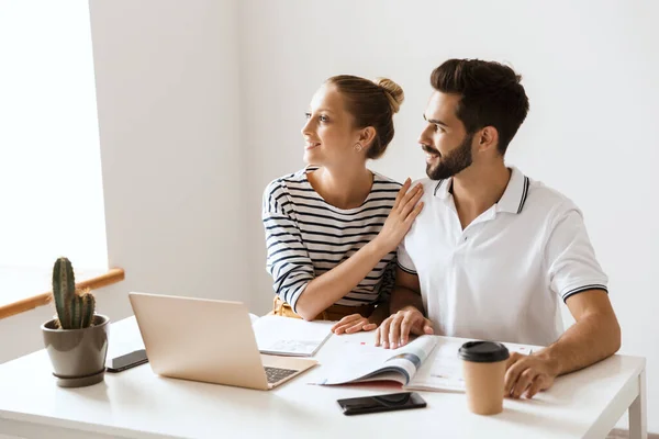 Piacevole allegro giovane coppia amorevole amici uomo e donna seduti al tavolo al chiuso utilizzando computer portatile studiare con notebook guardando la finestra . — Foto Stock