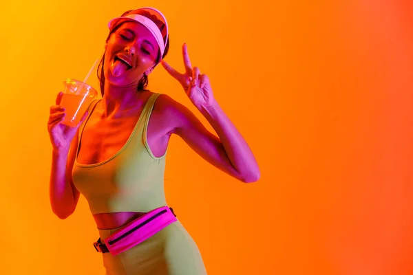 Känslomässig optimistisk elegant flicka i sport livsstil kläder och transparent hatt dricka juice cocktail isolerad över orange bakgrund med neon ljus. — Stockfoto
