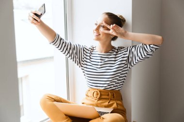 Neşeli genç pozitif kadın pencerenin yanına oturmuş dizüstü bilgisayar kullanıyor cep telefonuyla selfie çekiyor..