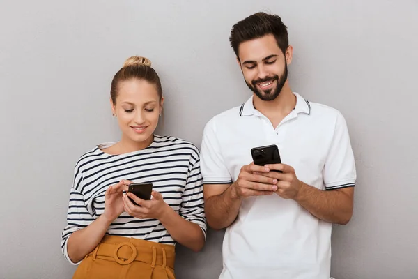 Blij jong positief liefdevol paar vrienden man en vrouw poseren binnen geïsoleerd over grijze achtergrond met behulp van mobiele telefoons. — Stockfoto