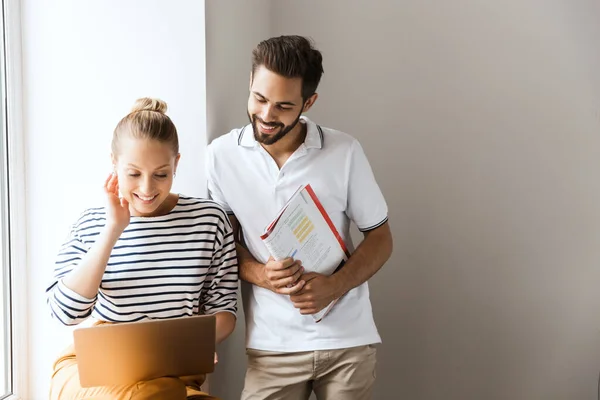 Jovem alegre feliz amoroso casal amigos homem e mulher perto da janela dentro de casa conversando uns com os outros segurando livros usando computador portátil . — Fotografia de Stock