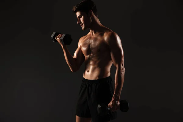 Concentrated handsome young strong sportsman posing isolated over black wall background holding dumbbells. — Stock Photo, Image