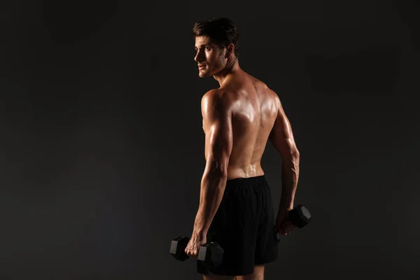 Concentrado guapo joven deportista fuerte posando aislado sobre fondo negro de la pared sosteniendo pesas . — Foto de Stock