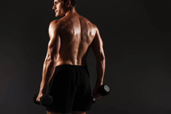 Concentrado guapo joven deportista fuerte posando aislado sobre fondo negro de la pared sosteniendo pesas . — Foto de Stock