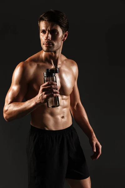 Concentrado bonito jovem forte desportista posando isolado sobre preto parede fundo garrafa com água . — Fotografia de Stock