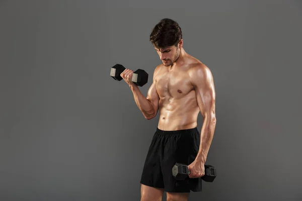 Guapo joven deportista fuerte posando aislado sobre fondo gris de la pared hacer ejercicios con pesas para los brazos . — Foto de Stock