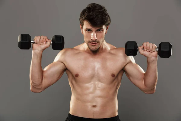 Guapo joven deportista fuerte posando aislado sobre fondo gris de la pared hacer ejercicios con pesas para los brazos . — Foto de Stock