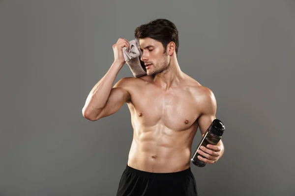 Tired handsome young strong sportsman isolated over grey wall background holding towel and bottle with water. — Stock Photo, Image