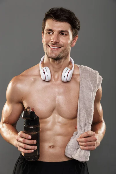 Glad leende stilig ung stark idrottsman isolerad över grå vägg bakgrund med hörlurar på halsen håller handduk och flaska med vatten. — Stockfoto