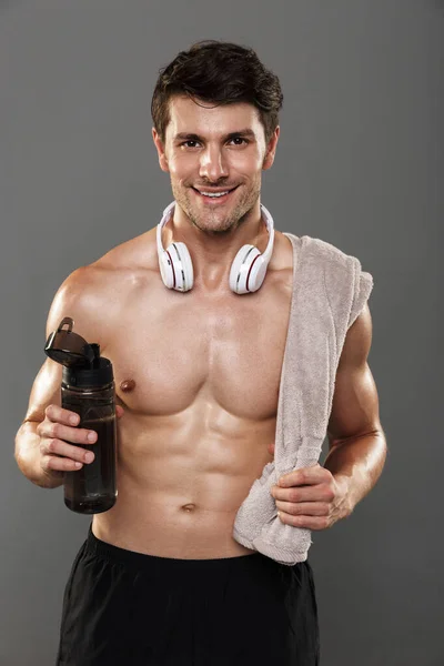 Happy smiling handsome young strong sportsman isolated over grey wall background with headphones on neck holding towel and bottle with water. — Stock Photo, Image