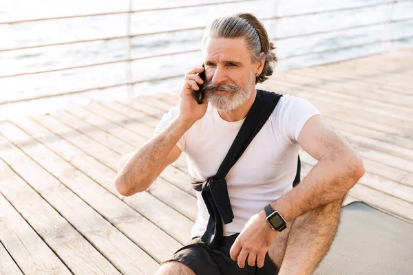 Image of pleased elderly man talking on cellphone while sitting on mat — Stock Photo, Image