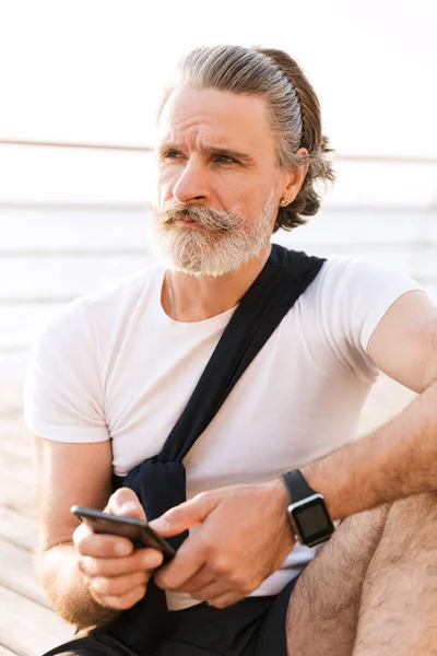Зображення вдумливого літнього чоловіка, який друкує на мобільному телефоні, сидячи — стокове фото