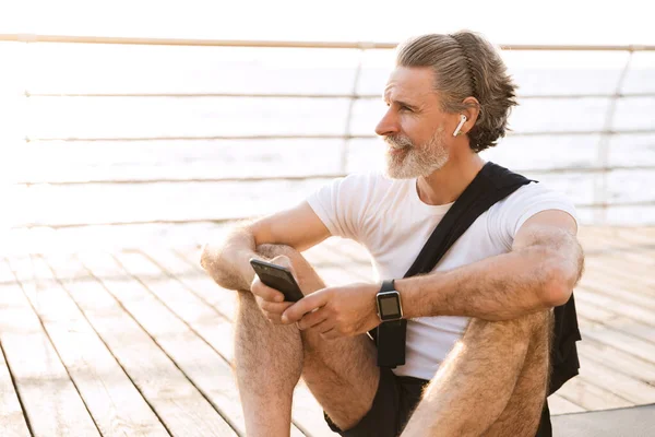 Εικόνα του ικανοποιημένου ηλικιωμένου άνδρα που χρησιμοποιεί το ακουστικό και το κινητό του ενώ κάθεται — Φωτογραφία Αρχείου