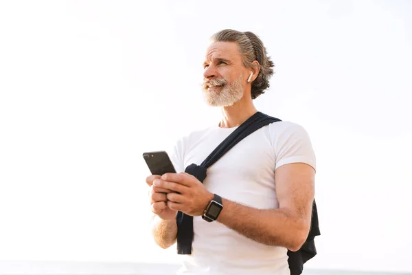 Immagine di vecchio compiaciuto utilizzando auricolare e cellulare mentre si lavora fuori — Foto Stock