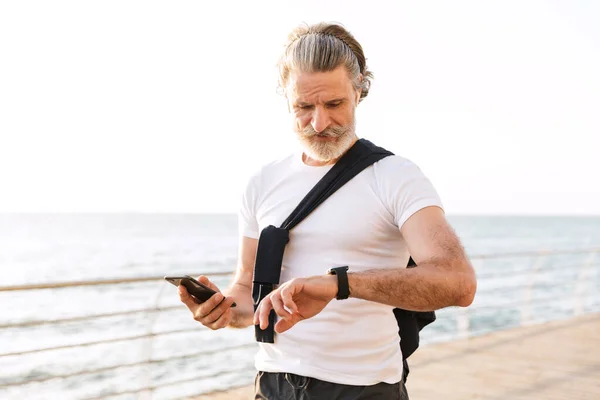 Immagine di uomo anziano soddisfatto utilizzando smartwatch e cellulare — Foto Stock