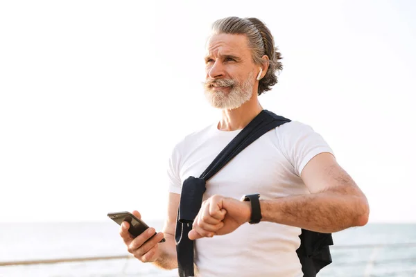 Obraz zadowolonego starszego mężczyzny korzystającego ze smartwatcha i telefonu komórkowego — Zdjęcie stockowe