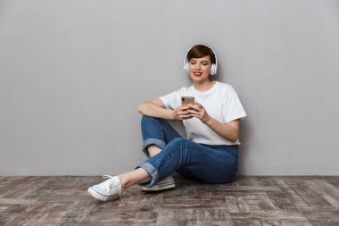 Yerde oturan cep telefonu ve kulaklık kullanan bir kadın resmi