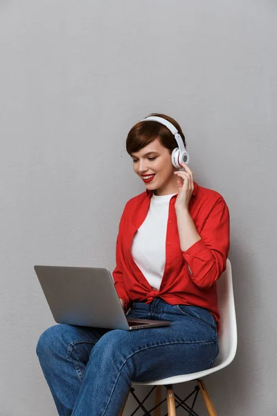 Image of pleased woman in headphones sitting on chair with lapto — Stok fotoğraf