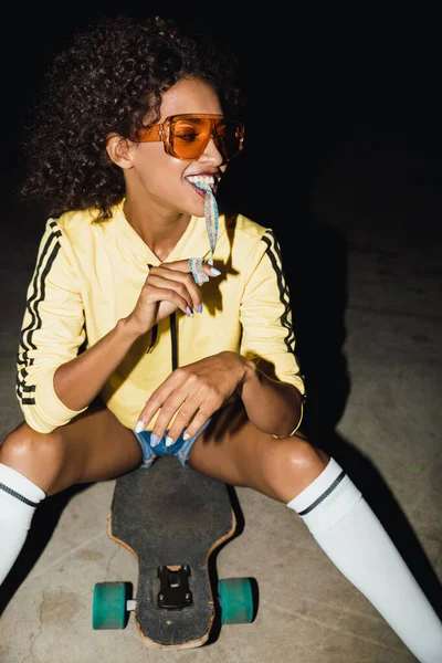 Imagem da menina americana africana comendo doces longos no skate um — Fotografia de Stock