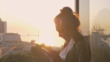 Balkonda dinlenirken akıllı telefonda gülümseyen ve sohbet eden neşeli esmer kadının yan görüntüsü.