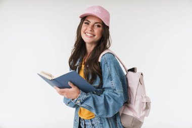 Sırt çantalı gülümseyen mutlu öğrenci beyaz duvar arkasına izole edilmiş ve elinde kitap tutuyor..