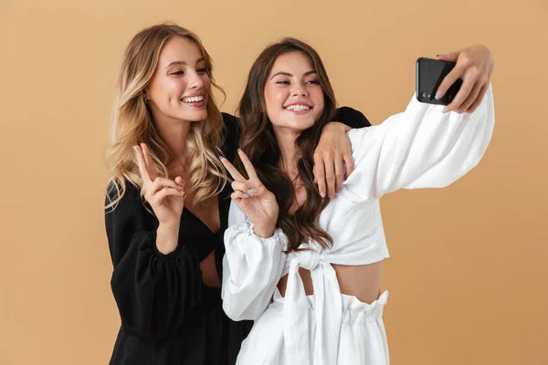 Retrato de dos mujeres sonrientes usando un smartphone mientras hacen gestos de paz —  Fotos de Stock