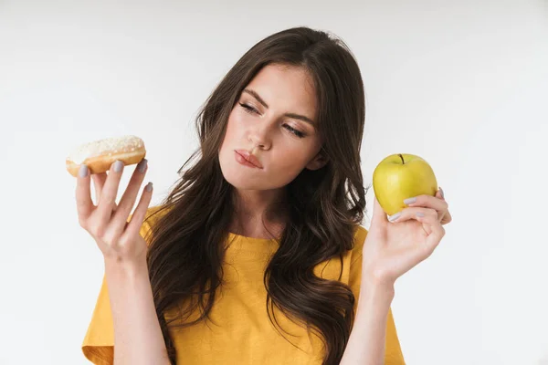 年轻女子站在白墙的背景上，在甜甜圈和健康苹果之间做出选择. — 图库照片