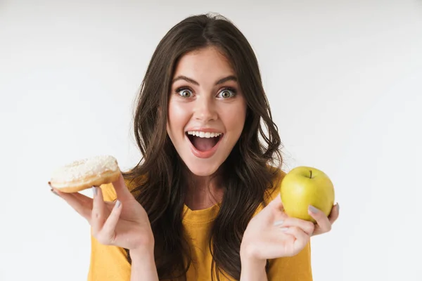 Fröhliche junge Frau posiert isoliert über weißem Wandhintergrund mit Donuts Süßigkeiten und gesundem Apfel. — Stockfoto