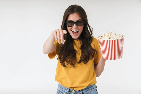 Lachende jonge vrouw geïsoleerd over witte muur achtergrond in 3D-bril eten popcorn horloge film wijzend naar u. — Stockfoto