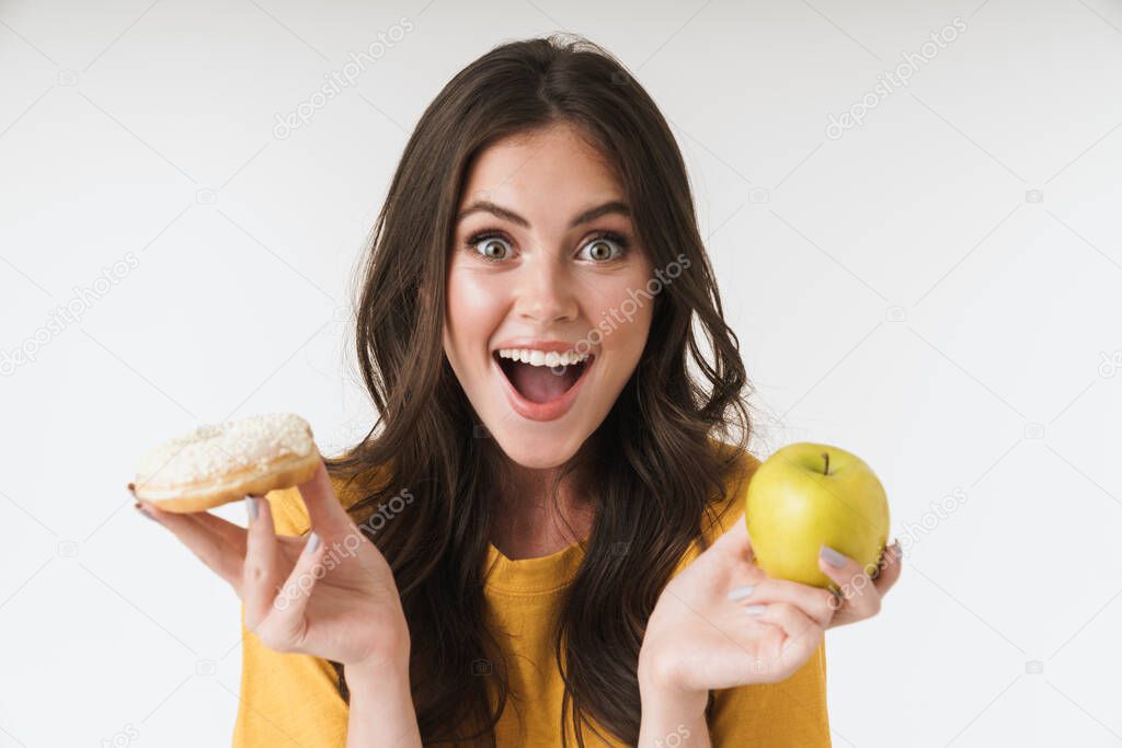 Cheery young woman posing isolated over white wall background holding donuts sweeties and healthy apple.