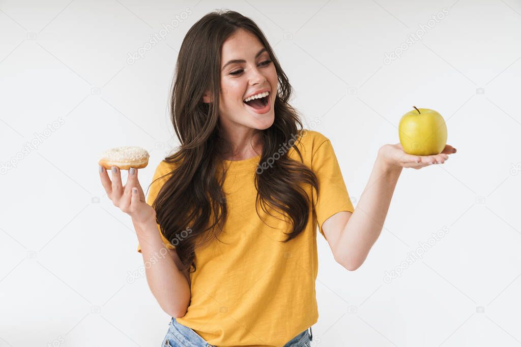 Smiling optimistic cheery young woman posing isolated over white wall background holding donuts sweeties and healthy apple.