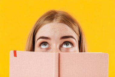 Sarı duvar arkaplanından izole edilmiş kız yüzünü defterle kapatıyor..