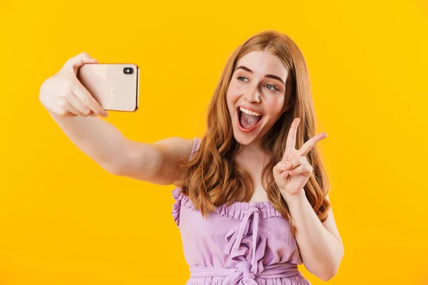 Veselý potěšené dívka izolované přes žlutou zeď pozadí vzít selfie mobilním telefonem ukazující mír gesto. — Stock fotografie