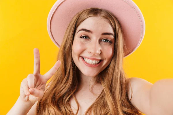 Happy girl isolated over yellow wall background wearing hat take selfie by camera with peace gesture. — ストック写真