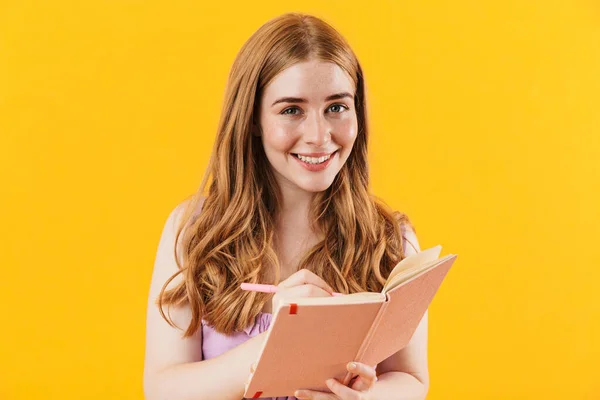 Smiling girl isolated over yellow wall background writing notes in notebook. — Zdjęcie stockowe