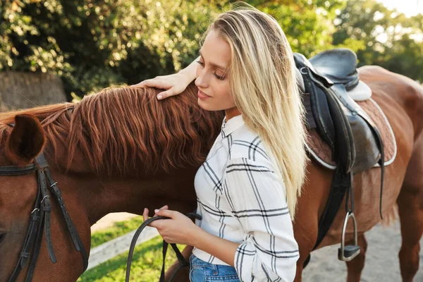 Bela sorridente jovem loira acariciando um cavalo no estábulo — Fotografia de Stock