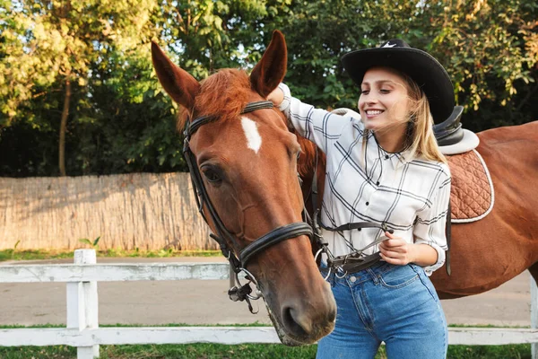 Hermosa joven rubia sonriente acariciando un caballo en el establo —  Fotos de Stock