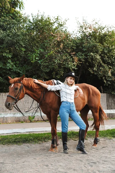 Beautiful smiling young blonde woman petting a horse at the stable — Stock Photo, Image