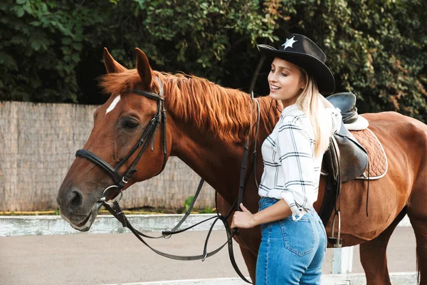 Bela sorridente jovem loira acariciando um cavalo no estábulo — Fotografia de Stock