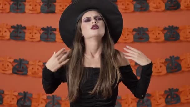 身穿黑色长袍的不快乐的年轻女巫厌倦了 她的胳膊交叉在橙色南瓜墙上 — 图库视频影像