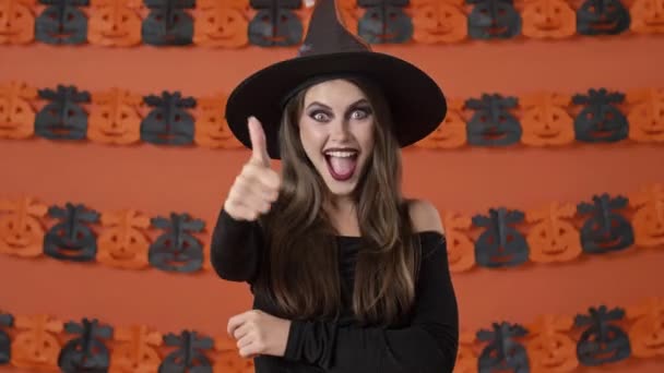 Atraktivní veselá mladá čarodějnice v černém halloween kostým dělat palec nahoru a ok gesto přes oranžové dýně zeď