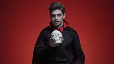 Siyah cadılar bayramı kostümlü Drakula adam kırmızı duvarın üzerinde insan kafatasını ve kükremesini gösteriyor.