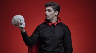 Siyah cadılar bayramı kostümlü korkunç vampir adam elinde insan kafatası tutuyor ve kırmızı duvarın üzerinde izole edilmiş bir şey anlatıyor.