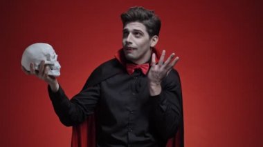 Siyah cadılar bayramı kostümlü korkunç vampir adam elinde insan kafatasını tutuyor ve onunla kırmızı duvarın üzerinde konuşuyordu.