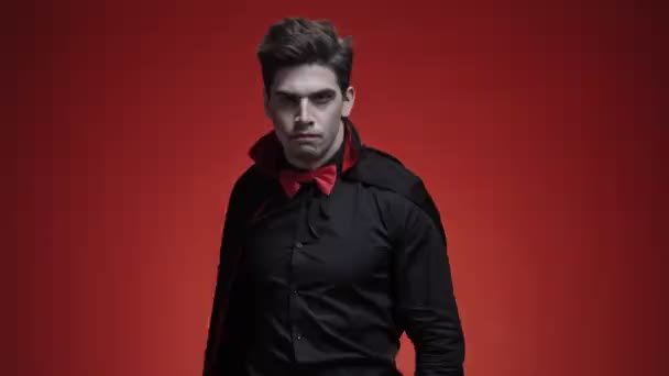 吸血鬼男とともに血と牙で黒ハロウィン衣装ラップで彼のクローク隔離された上の赤い壁 — ストック動画